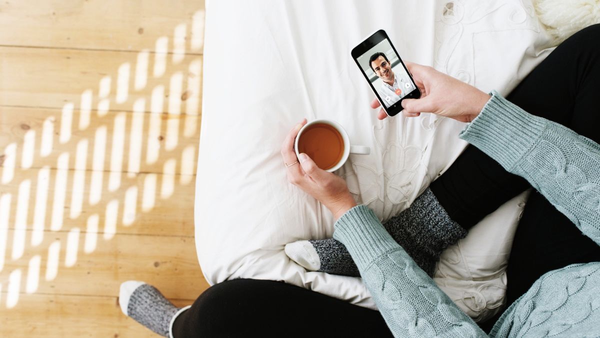 Eine Frau sitzt zuhause mit einer Tasse Kaffee und nutzt das Smartphone für eine Onlinesprechstunde der TeleClinik