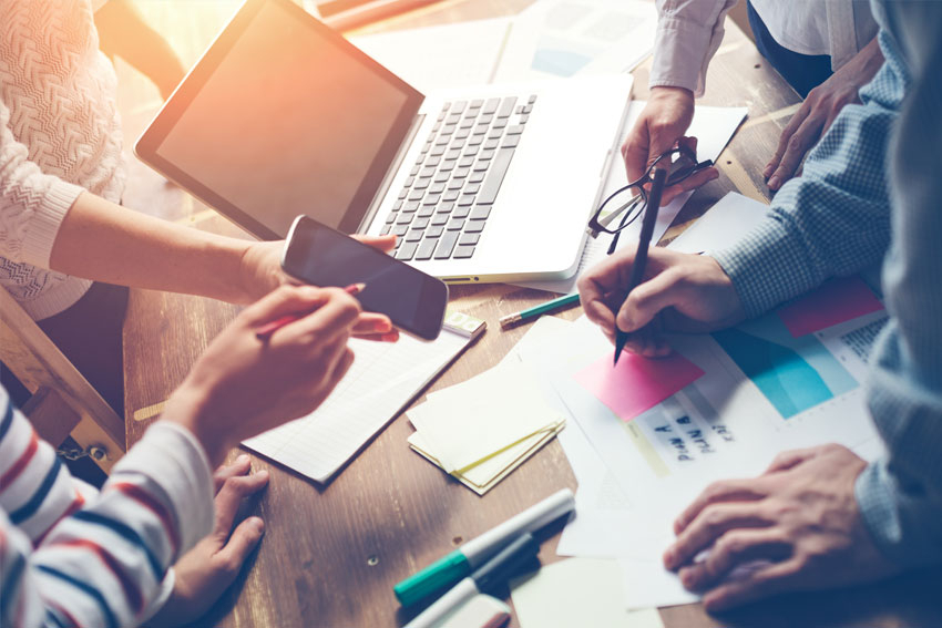 Ausschnitt Personen am Tisch mit Laptop, Stift, Handy, Papier