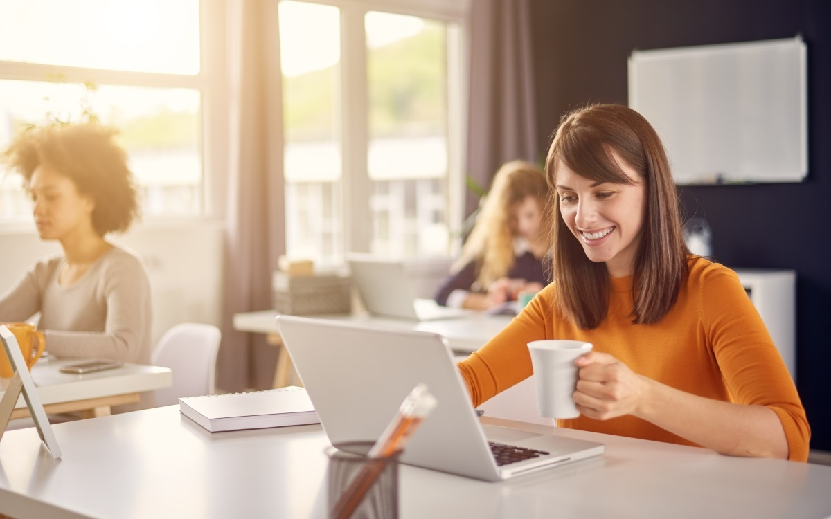 Frau mit Tasse in der Hand arbeitet am Laptop.
