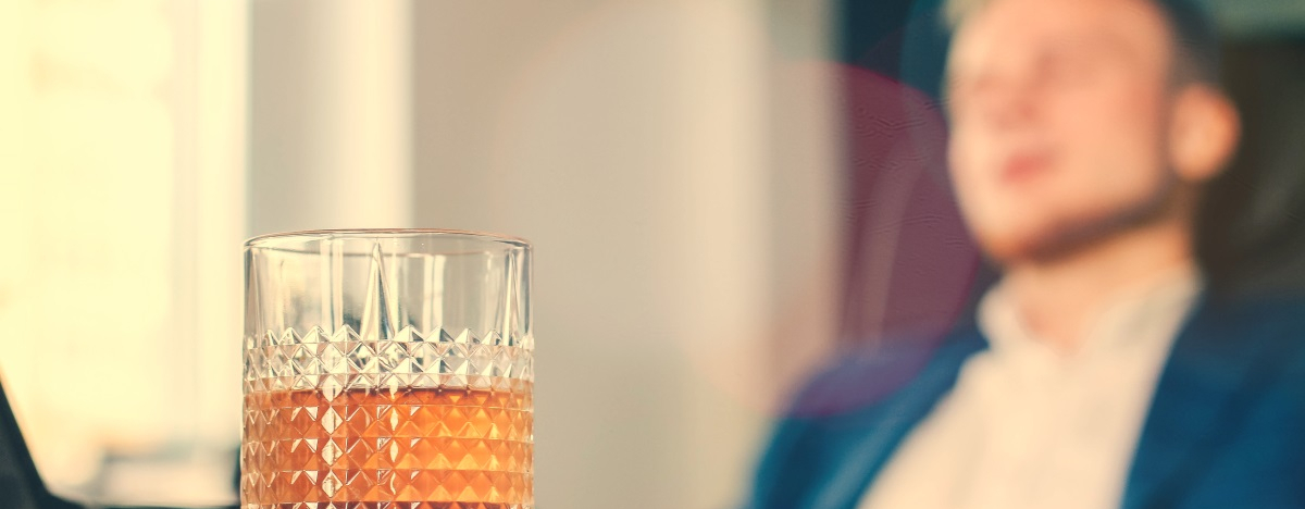 Ein Glas Whisky, im Hintergrund ein schlafender Mann