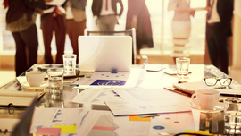 Unterlagen auf Konferenztisch und stehende Teilnehmer im Hintergrund