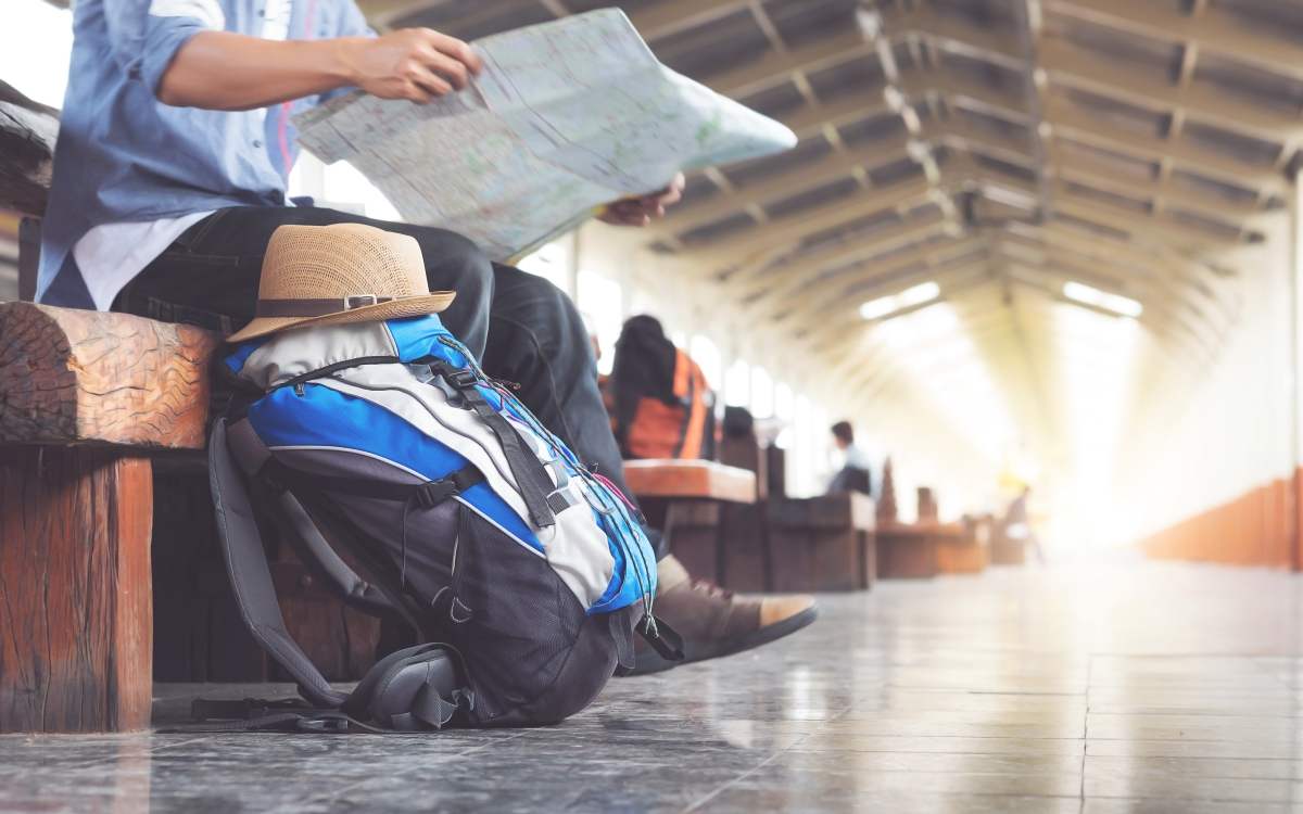 Mann mit Rucksack sitzt am Bahnhof und liest Zeitung, w&auml;hrend er auf einen Zug wartet