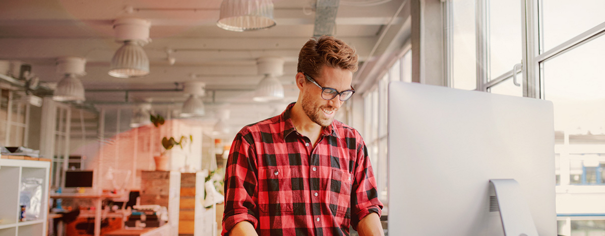 Junger Mann steht lachend am Schreibtisch, vor ihm der Monitor