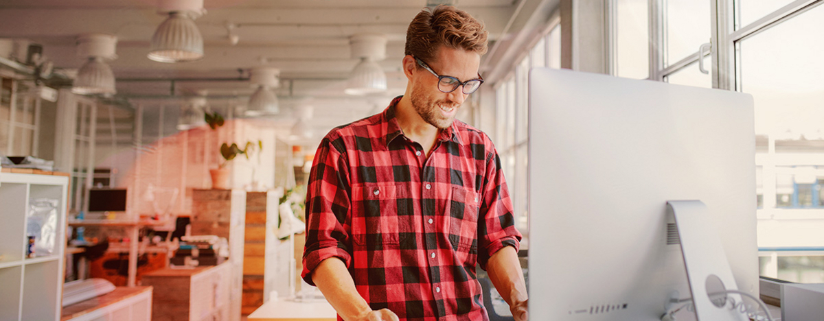 Junger Mann steht lachend am Schreibtisch, vor ihm der Monitor